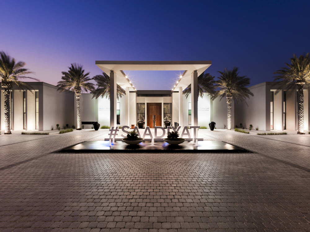 Saadiyat Beach Club at dusk. The modern and white palm tree lined building has "#SAADIYAT" in front of it
